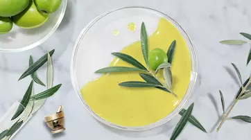 A clear dish with a smooth, vibrant green olive oil or dressing, accompanied by fresh olives and olive leaves, set against a marble background.