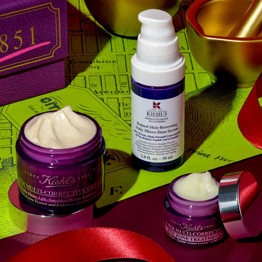 Three Kiehl's Super Multi- Corrective products—a cream jar, an eye cream, and a serum bottle—are arranged on a red ribbon, beside a purple box and gold festive decorations, against a lime green map- patterned background.