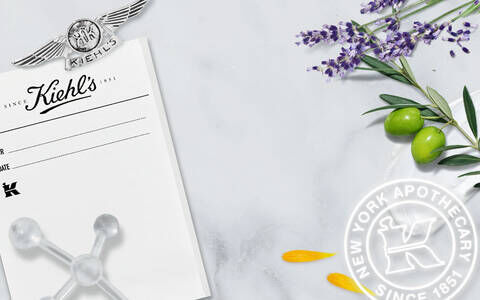 A marble background featuring a vintage black-and-white photo of Kiehl's pharmacy staff, along with lavender, olives, and decorative elements, emphasizing Kiehl's history as a New York apothecary since 1851.