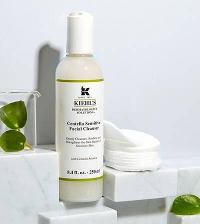 Čistač lica Kiehl's Centella Sensitive, u staklenoj boci, sa pamukom za negu osetljive kože na marmernoj podlozi.