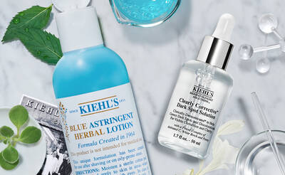 A flat lay of Kiehl's skincare products, featuring a blue astringent herbal lotion and a clear dark spot solution, surrounded by vibrant leaves and a blue gel.