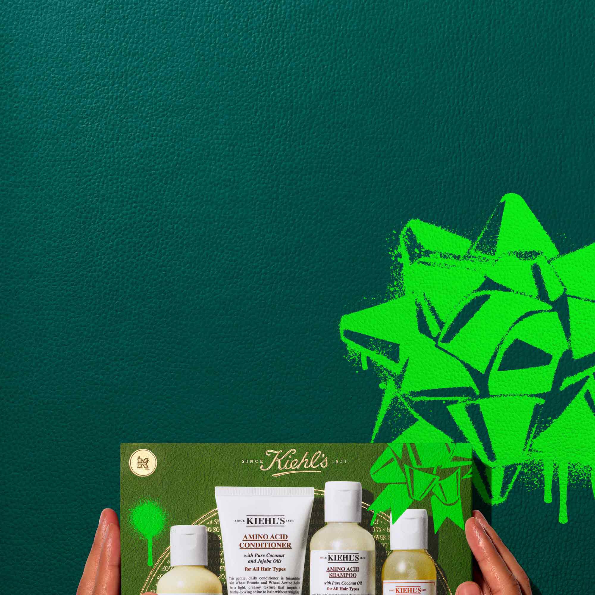 A graphic, set against a dark green textured background, features four products. The top row displays: a bottle of "KIEHL’S CREME DE CORPS," a tube of "KIEHL’S AMINO ACID CONDITIONER," a bottle of "KIEHL’S AMINO ACID SHAMPOO," and a bottle of "KIEHL’S BATH AND SHOWER LIQUID BODY CLEANSER GRAPEFRUIT." Below them, a green Kiehl’s gift box showcases the same four products neatly arranged inside. The gift box has bright green graphic accent.