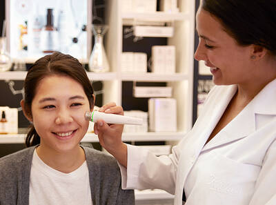 A skincare professional uses a device on a smiling client's face in a bright, modern retail space.
