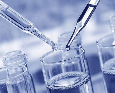 A dropper is releasing liquid into a glass container, surrounded by other glass containers filled with liquid, in a laboratory setting.