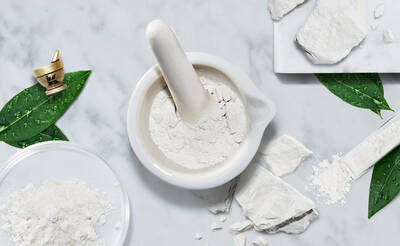 A white mortar and pestle surrounded by loose white powder, broken pieces of powdery substance, and green leaves on a light marble surface.