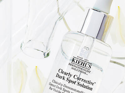 A bottle of Kiehl's Clearly Corrective Dark Spot Solution surrounded by white petals and a glass container.