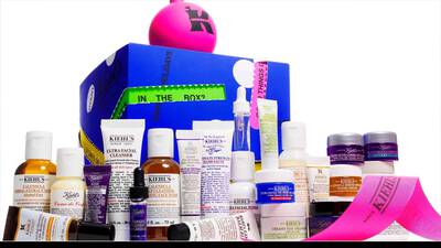 A colorful display of Kiehl's skincare products, including various creams, cleansers, and serums, arranged around a bright pink box with holiday-themed packaging.