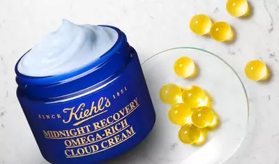 A blue jar of Kiehl's Midnight Recovery Omega-Rich Cloud Cream with a light blue cream on top, next to a clear plate containing yellow capsules.
